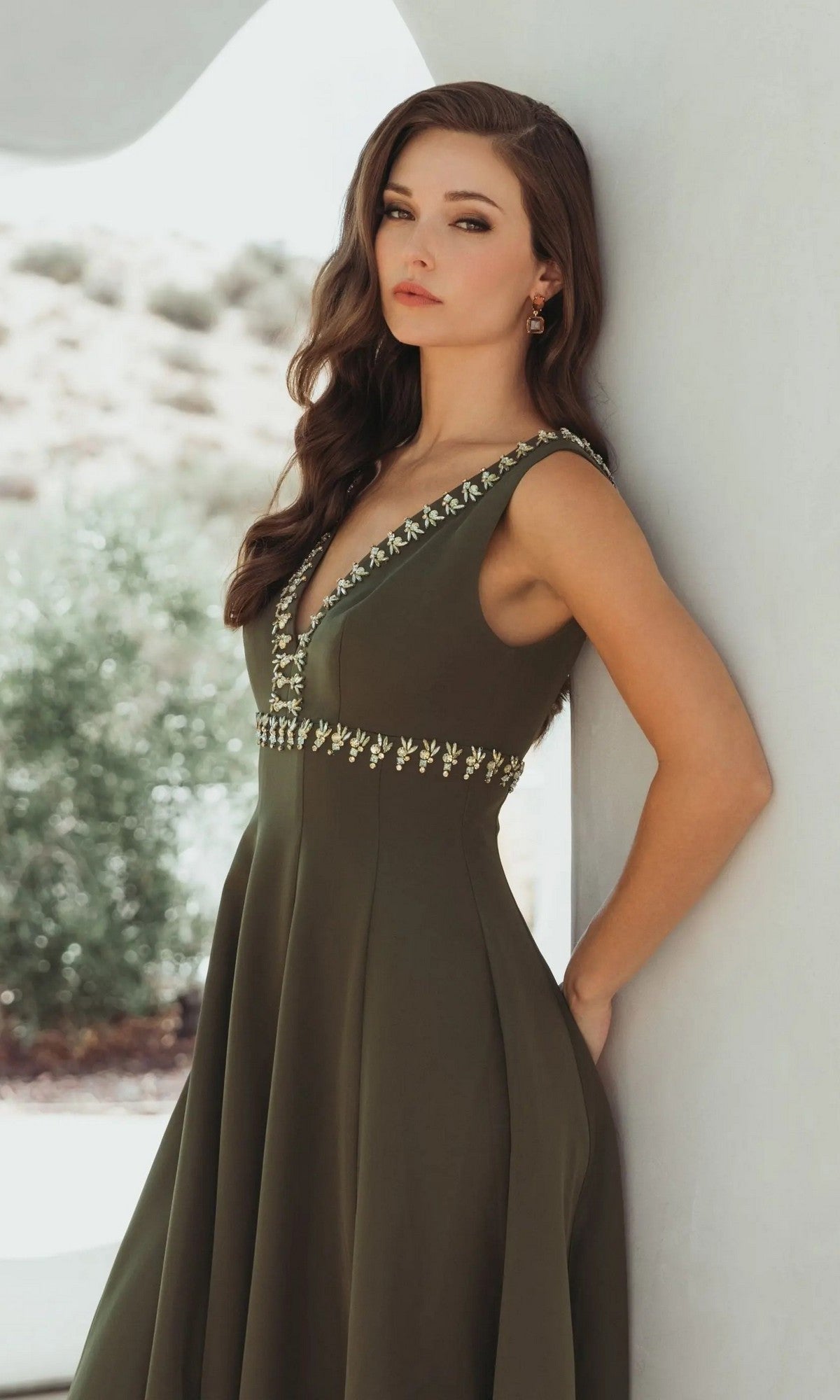 A woman with long brown hair wears the Mac Duggal 21159, an olive green sleeveless midi dress with embroidered details—an elegant choice for a wedding guest.